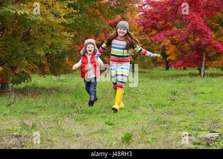 Ein junger Junge und Mädchen tragen Gummistiefel und bunte Kleidung laufen über Ein Feld Halten Sie Hände mit Bäumen in hellen Herbstfarben im... Stockfoto