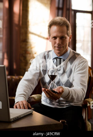 Psychiater Mann Lächeln für die Kamera und halten Sanduhr in der hand während des Wartens auf Patienten in seinem Büro. Porträt der schönen Psychiater Mann aussehen Stockfoto