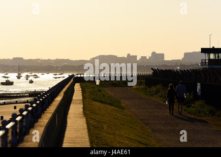 Ein Paar geht Hand in Hand in Hand in Richtung einer niedrigen Sonne entlang eines Uferpfades mit der Silhouette der Stadt Southend im Hintergrund, Boote und Radfahrer Stockfoto