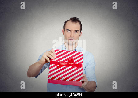 Closeup Portrait mürrischer unglücklich aufgeregt Mann halten rote Geschenkbox sehr unzufrieden mit dem, was er erhalten, Ekel auf grauem Hintergrund Gesicht isoliert. Negati Stockfoto