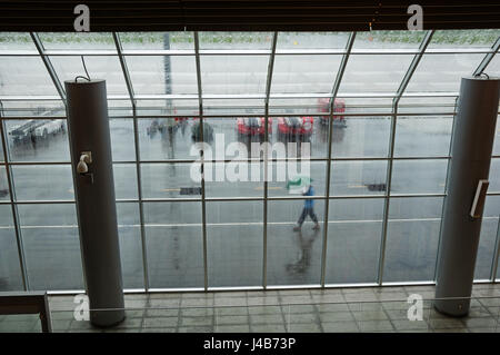Blick durch ein Fenster der Flughafen an einem regnerischen Tag mit einer Person zu Fuß von außen mit Regenschirm Stockfoto