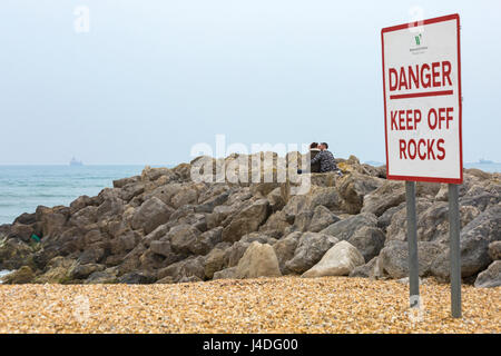 Schild mit der Aufschrift „Danger keep off rocks“ (Gefahr, nicht auf Steinen halten), auf dem ein Paar im Mai in Weymouth, Dorset UK, sitzt und sich küsst Stockfoto