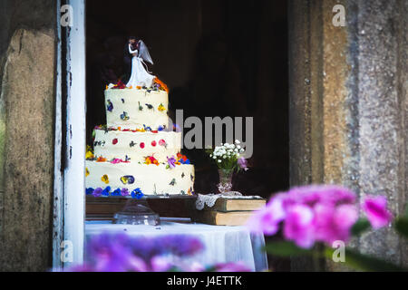 Hübsche Hochzeitstorte mit weißer Glasur und Blumen garniert mit Figuren der Braut und des Bräutigams. Rosa und lila Blumen im Vordergrund unscharf zurück Stockfoto