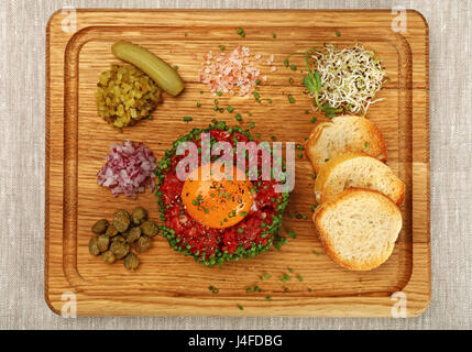 Eine Portion rohes Hackfleisch Fleisch Tartar Steak mit Eigelb, Zwiebeln, Gurken, Kapern, Salz und Toast serviert am Holzbrett, Nahaufnahme, erhöhte Stockfoto