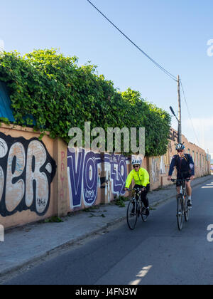 Zwei Touren Radfahrer ihr Fahrrad entlang einer Straße, flankiert von einer Wand bedeckt in Graffiti. Stockfoto