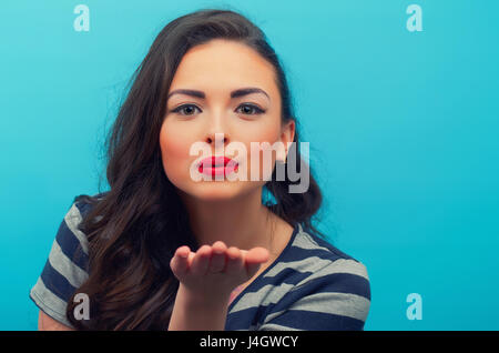 Porträt des schönen lächelnden Mädchens, einen Luft-Kuss auf dem blauen Hintergrund sendet. Junge weibliche Modell. Stockfoto