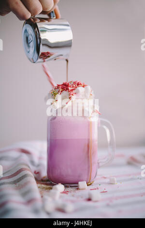 Frau gießt Kaffee in stilisierten Einmachglas Tasse farbige Milch mit Sahne, Eibisch und bunte Dekoration. Milch-Shake, Cocktaill, Frappuccino. Stockfoto