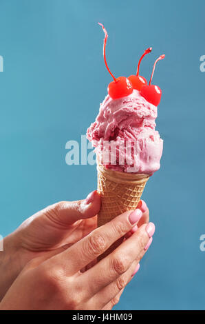 Weibliche Hände rosa Eistüte dekoriert mit Cocktail-Kirsche auf blauem Hintergrund Stockfoto