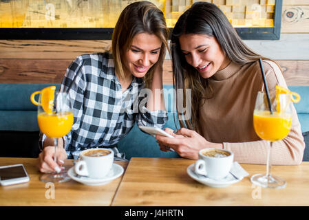 Zwei junge Freundinnen sitzen im Café, trinken Kaffee und Saft, etwas auf dem Handy zu sehen. Stockfoto