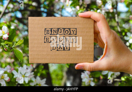 Frau Hand Holding Pappkarte mit Worten genießen Sie jeden Tag von schwarzen Alphabet Stempel gegenüber blühenden Baum Hintergrund gemacht. Stockfoto