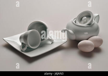 Gerichte aus chinesischem Porzellan weiß: eine Teekanne mit einem angehobenen Deckel, zwei Tassen, eine Untertasse, neben dem Kessel liegen zwei rosa Hühnereier. Stockfoto