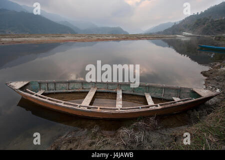 Großen alten Holzboot über das Ufer am See Feva, spiegelt das Wasser eine düstere grauen Himmel im Hintergrund des Berges, Pokhara, Nepal. Stockfoto