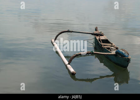 Kleinen alten balinesischen Holzboot, blaue Farbe auf der Seite, gebrochene Sitze auf der linken Seite ein großes Gegengewicht steht auf dem Wasser, das weiße Clo widerspiegelt Stockfoto