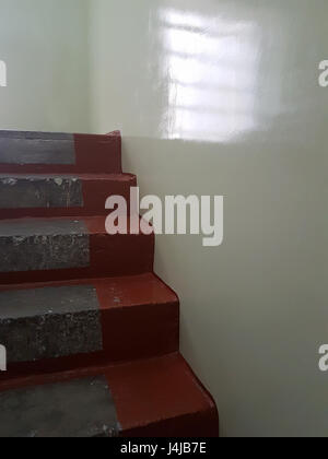 Vintage Zement Treppen an den Seiten in Burgund Farbe bemalt, leicht graue Wand und aus dem Fenster an der Wand, Treppe alte Haus innen reflektiert Stockfoto