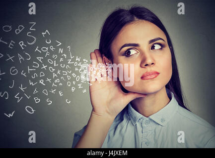 Frau hält ihre Hand in der Nähe von Ohr und hört aufmerksam Alphabet Buchstaben fliegen auf graue Wand Hintergrund isoliert Stockfoto