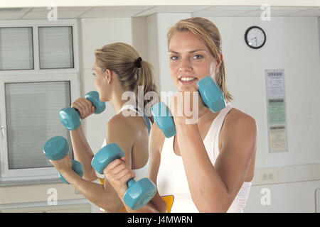 Fitness-Studio, Frau, jung, Ständer, Hanteln, Fitness üben, an der Seite, halb Porträt-Fitness-Raum, Sportlerin, Blond, Sportkleidung, sportlich straffen, Sport, Hobby, Freizeit, Fitness, Gesundheit, Bewegung, Praxis, Muskulatur, Muskeln, Arme, Bizeps, Arm Muskulatur, Armmuskeln, Dehnung, Muskelaufbau, Training, Zug, Hantel Training, Gewichte, kurz Hanteln, Position, Position, Lächeln, view Kamera, innen, Hintergrund, Reflektor, Reflexion Stockfoto