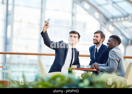 Gruppe junger lächelnd Geschäftsleute nehmen Selfie am Tisch während der Sitzung in der Glashalle moderne Bürogebäude Stockfoto