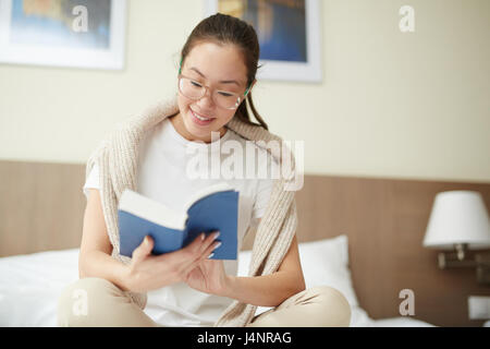 Porträt von lächelnden Asiatin mit Brille Buch sitzt auf Bett tagsüber Stockfoto