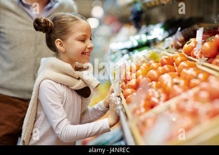 Seite Ansicht Porträt der niedliche kleine Mädchen lächelnd schiefen über Gemüsetheke mit frischen reifen Tomaten und anderes Gemüse Stockfoto