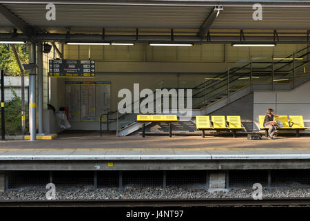 Eine Frau sitzt auf einer hellen gelben Kunststoff Bank während des Wartens auf den nächsten Zug am Bahnhof Liverpool South Parkway in South Liverpool. Stockfoto