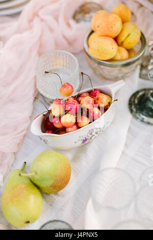 Picknick, Essen, Urlaub Konzept - leckere frische rote und gelbe Kirschen, Birnen, Aprikosen auf dekorierten Tisch für das Picknick, Früchte Teller und Geschirr, veganes Essen, weißes Tischtuch, reinigen Sie frisches Obst. Stockfoto