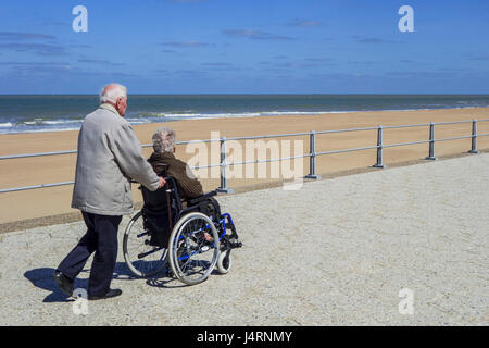 Pensionierter Mann unter deaktiviert ältere Frau im Rollstuhl für einen Spaziergang auf der Promenade entlang der Küste an einem kalten, sonnigen Tag im Frühling Stockfoto