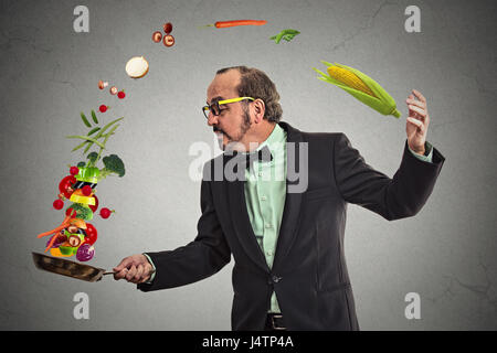 Businessman Kochen Gemüse mit einer Pfanne auf graue Wand Hintergrund isoliert. Positive Gesicht Ausdruck Emotion. Stockfoto