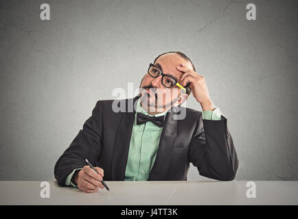Verwirrt Lehrer sitzen am Tisch, kratzte sich am Kopf denken was für Idee, isolierte Büro graue Wand Hintergrund mit textfreiraum zu schreiben. Menschliches Gesicht exp Stockfoto
