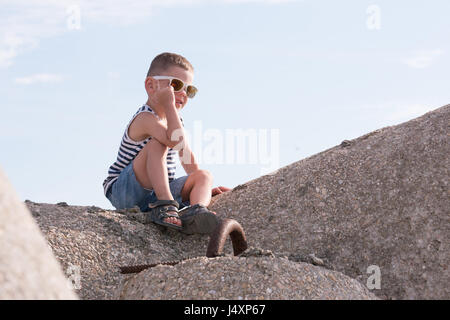 fröhliche kleine Junge in Sonnenbrillen, Sailor Streifen Weste und Shorts sitzen auf einem Wellenbrecher lächelnd Stockfoto