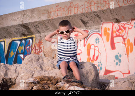 Schöne kleine Junge in trendigen Sonnenbrillen und Seemann Weste sitzen auf Stein vor Wand mit graffiti Stockfoto