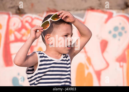 kleiner Junge mit weiße Sonnenbrille und Streifen Matrosenhemd auf gelb und rosa Graffiti Wand Hintergrund erstaunt Stockfoto