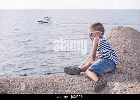 Kleiner Junge mit einer Sonnenbrille und gestreifte Weste auf konkrete Wellenbrecher liegt direkt am Meer schwimmende Boot sitzen Stockfoto