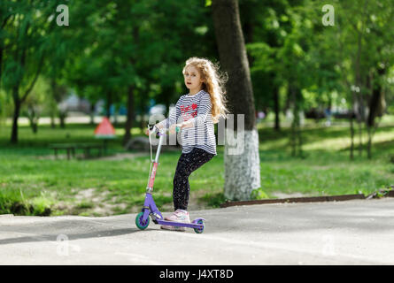 Kleine blonde Mädchen fahren die Roller in den Park. Stockfoto