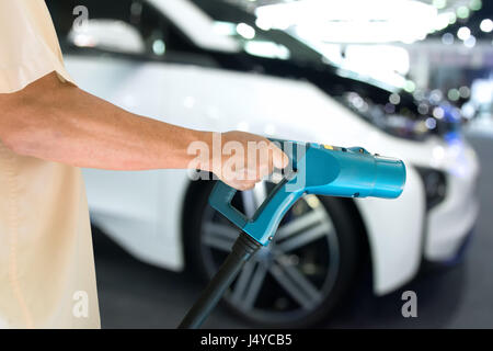 Mann Hand halten und laden Elektro-Auto mit dem Netzteil, Kabel eingesteckt in mit Weichzeichnen Ev Auto Hintergrund Stockfoto