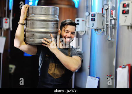 Mann mit einem Bart in Uniform mit einem Fass Bier in einer Brauerei mit Metallbehälter im Hintergrund arbeiten Stockfoto