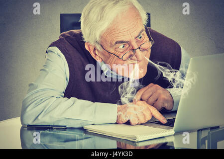 Älteren Mann mit Computer bläst Dampf aus Nase frustriert sitzen am Tisch, die isoliert auf graue Wand Hintergrund hervorgehoben. Senioren und Technik Stockfoto