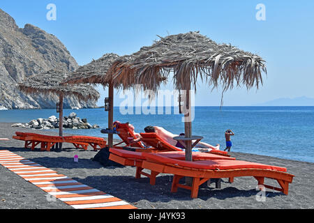 Touristischen ruht auf Sonnenliegen unter Stroh Sonnenschirmen in Perissa, Santorin, Kykladen, Griechenland Stockfoto