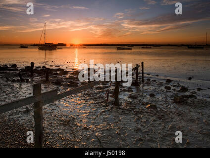 Eine helle orange Sonnenuntergang über der Mündung des Flusses Alde in Orford in Suffolk mit Silhouette Boote vor Anker im Fluss bei Ebbe und Wattenmeer Stockfoto