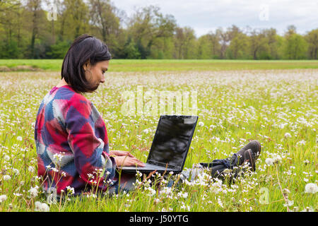 Kolumbianische Frau sitzt mit Laptop in Wiese mit blühenden Blumen Stockfoto