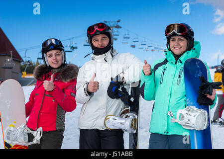 Jugendliche in Ski-Anzüge, Helme und Ski goggles Daumen nach oben, während mit stehen am Zaun in ein Skigebiet in Winterzeit snowboards Stockfoto