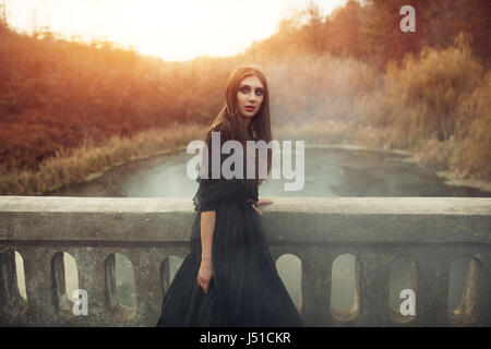 Junge attraktive Hexe im schwarzen Kleid zu Fuß auf die alte Brücke in schweren schwarzen Rauch. Dunkle Magie. Horror-Märchen. Stockfoto