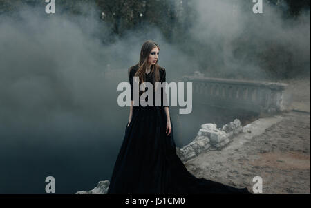 Junge attraktive Hexe im schwarzen Kleid zu Fuß auf die alte Brücke in schweren schwarzen Rauch. Dunkle Magie. Horror-Märchen. Stockfoto
