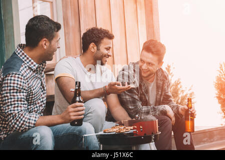 Lächelnde junge Freunde, Bier trinken und reden beim Grillen Fleisch im freien Stockfoto