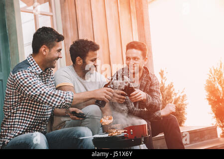 Lächelnde junge Männer Klirren Bierflaschen und Grillen von Fleisch auf Veranda Stockfoto