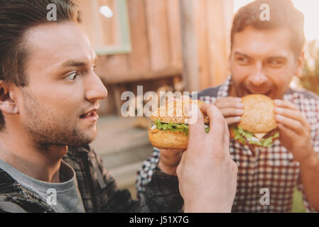 Emotionale junge Männer, die Gourmet-Hamburger im Freien essen Stockfoto