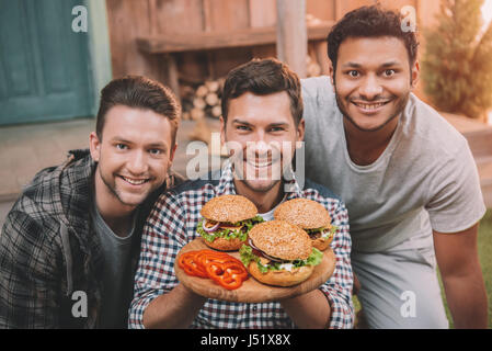 Drei glückliche junge Männer mit leckeren Burgern lächelnd in die Kamera Stockfoto