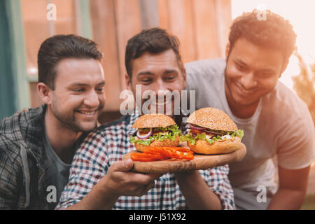 Drei lächelnde Männer betrachten frische hausgemachte Hamburger auf Holzbrett Stockfoto