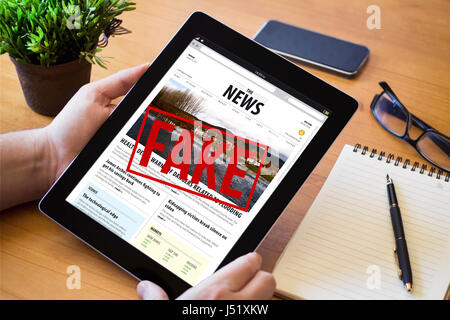 Hände eines Mannes Haltevorrichtung eine gefälschte Nachrichten über eine hölzerne Workspace-Tabelle. Alle Bildschirm-Grafiken bestehen. Stockfoto