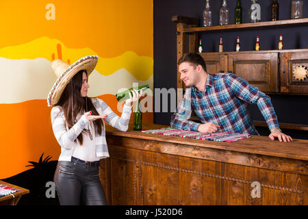Eine junge Frau in einen Sombrero hält ein Bier und stehen an der Bar neben der Barkeeper in einem karierten Hemd in einer mexikanischen bar Stockfoto