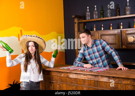 Eine junge Frau in einen Sombrero halten zwei Flaschen Bier und stehen an der Bar neben der Barkeeper in einem karierten Hemd in einer mexikanischen bar Stockfoto